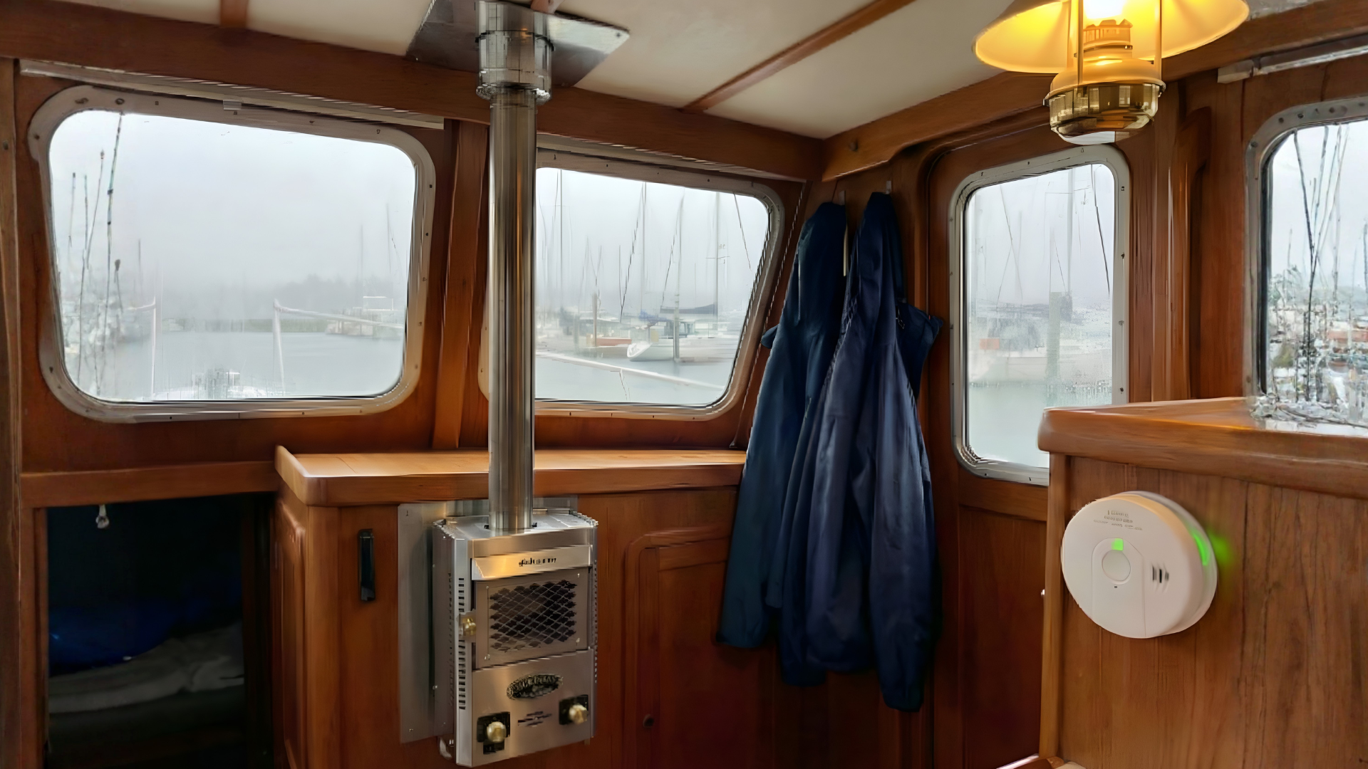 Interior of a wooden sailboat cabin featuring a stainless steel Dickinson Marine heater, wood-paneled walls, and views of a foggy marina through the windows