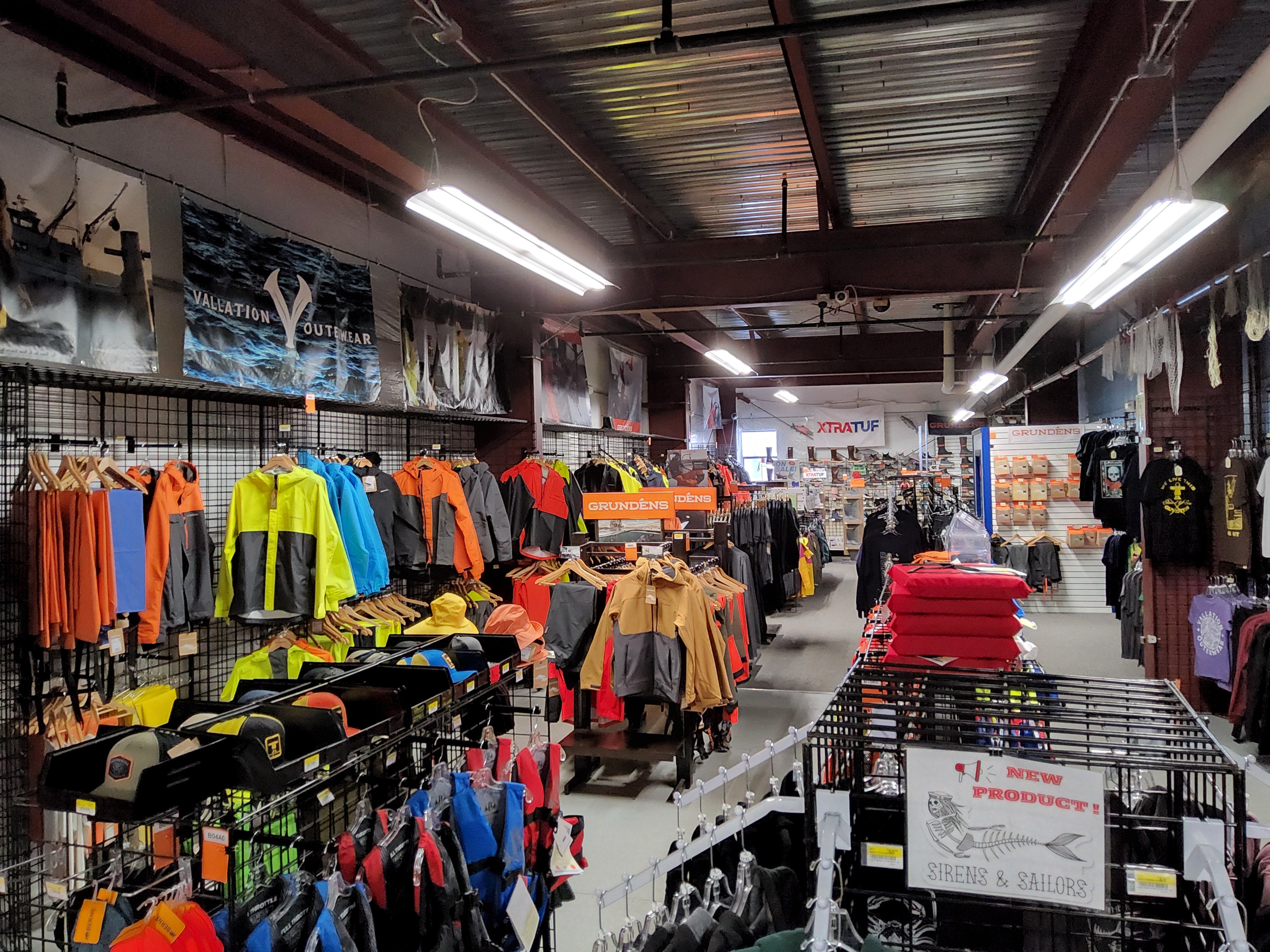 A retail store interior showing racks of outdoor and fishing apparel, including bright jackets, shirts, and hats, with wall displays and fluorescent lighting overhead.