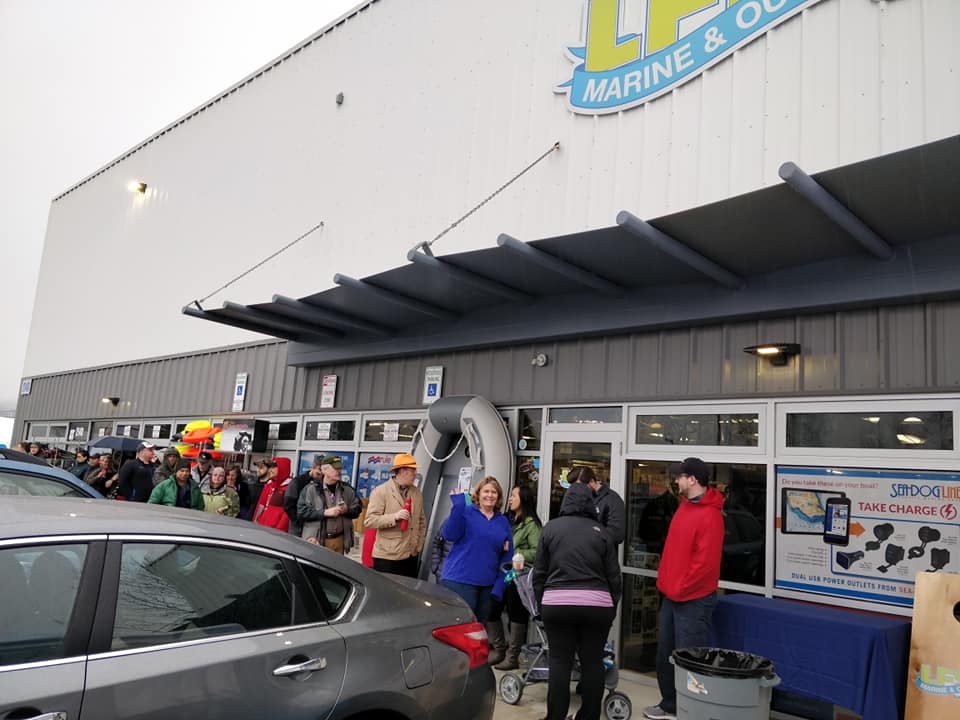 A crowd of people gathered outside the entrance of a large white warehouse-style store building under a dark awning. A blue and yellow sign on the building reads 'LP MARINE & OIL', and a gray sedan is visible in the foreground.