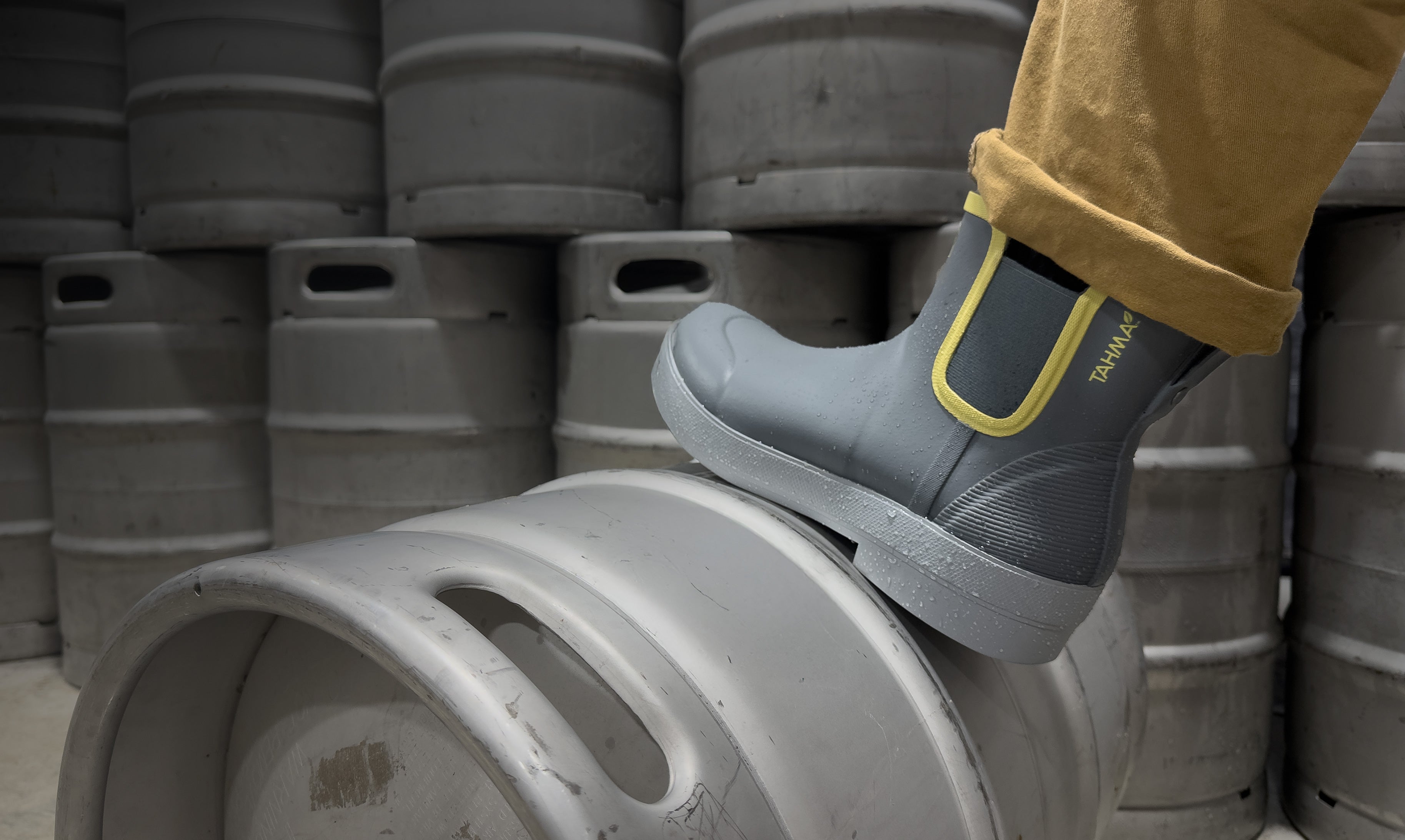 Person wearing gray rain boots with yellow soles standing on a barrel with stacked barrels in the background.