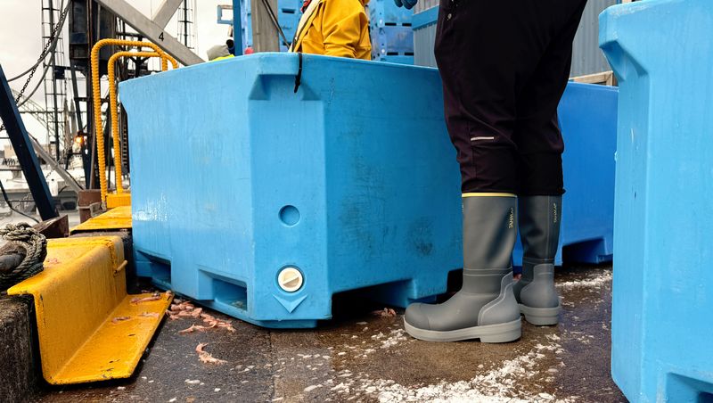 A close-up of a large blue plastic floating storage container on a work deck, with a person wearing dark pants and gray rubber Tahma boots standing beside it.